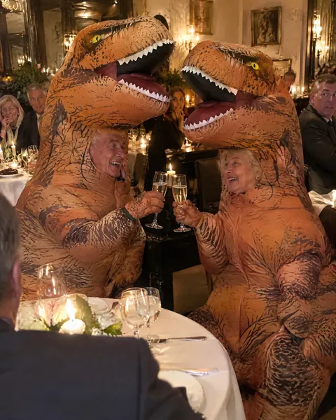 An older couple in matching inflatable T-rex costumes toasting at a candlelit anniversary dinner