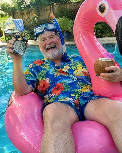 A retired man in a Hawaiian shirt on a flamingo pool float holding a smashed alarm clock