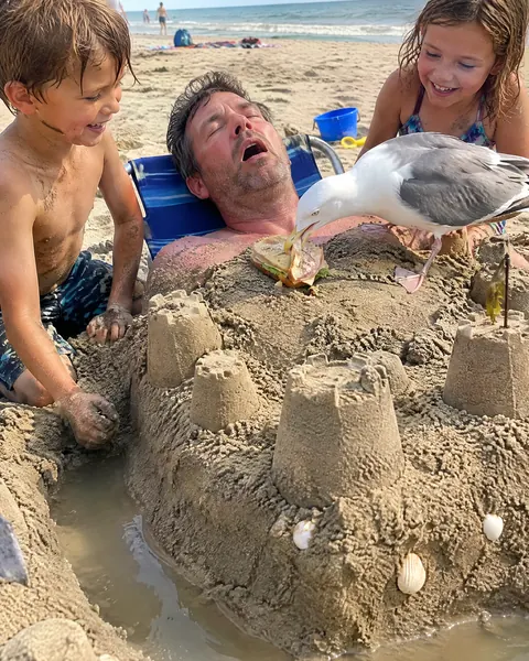 A dad asleep in a beach chair buried in a sandcastle while a seagull takes his sandwich